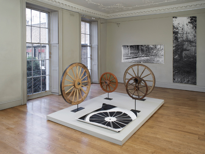 A Circle, image foreground:  Rupert Ackroyd + David Gates 'Cart Wheel Exercise' 2014, a group of 5 works as follows: (i) 112 cm dia, oak, elm, steel on steel base, (ii) 70 cm dia, perspex, oak, ash, elm, paint, steel on steel base, (iii) 111 cm dia, pine, chestnut, steel on steel base, (iv) 60 cm dia, pine, steel on steel base, (v) 106 × 120 cm, unique photogram on paper, base: 200 × 300 cm, mdf, pine, paint; background left: David Gates 'Standard Image #03' unique photograph 89 × 218 cm / 35 × 85.8 in (approx) 2014; background right:  David Gates 'Standard Image #10' unique photograph 292 × 89 cm / 114 × 25 in (approx) 2014; installation photography by Andy Keate